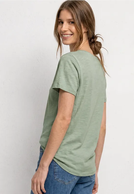 Seitlich abgebildete Frau mit grünem T-Shirt und blauen Jeans vor einem hellen Hintergrund.