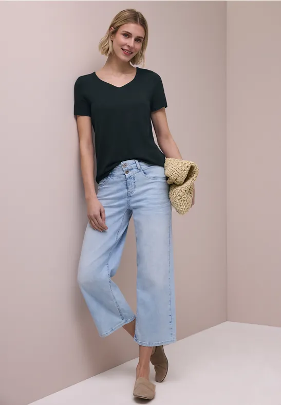 Vrouw in zwart T-shirt met lichtblauwe wijde jeans, strooien tas en beige sleehakken voor een neutrale achtergrond.