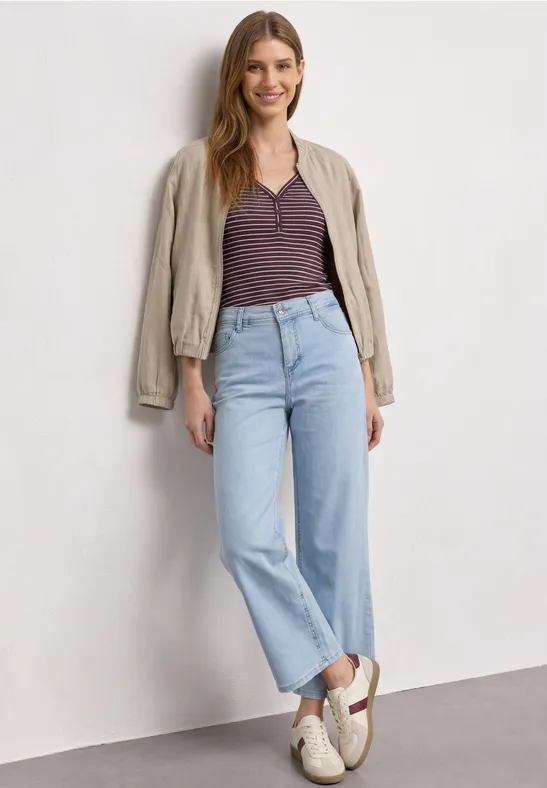 Eine Frau steht vor einer weißen Wand, trägt hellblaue Straight-Leg-Jeans, gestreiftes Shirt und beigen Blouson.
