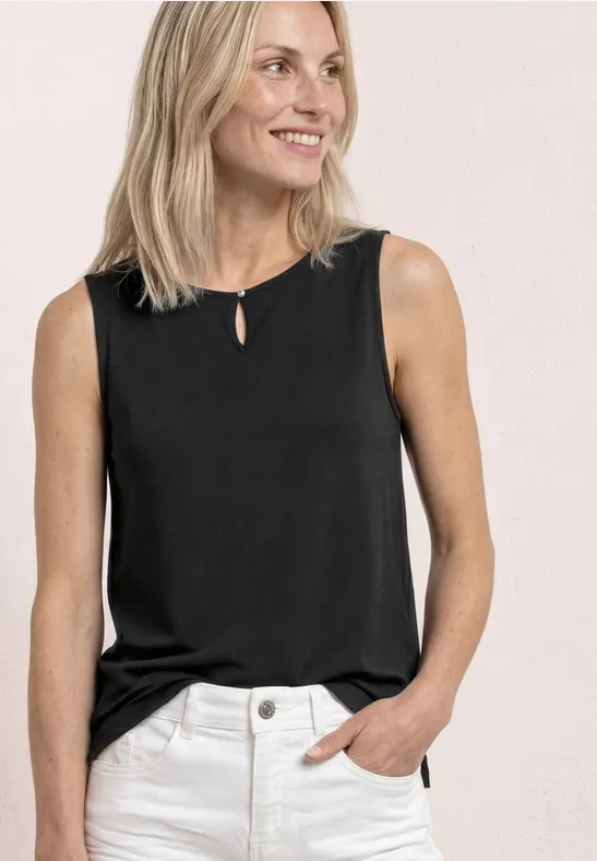 Vrouw draagt zwart mouwloos topje en lichte broek, hand in zak, tegen lichte achtergrond.