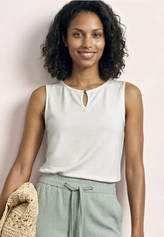 Vrouw in mouwloze witte top en lichtgroene broek, met een strooien tas in de hand.