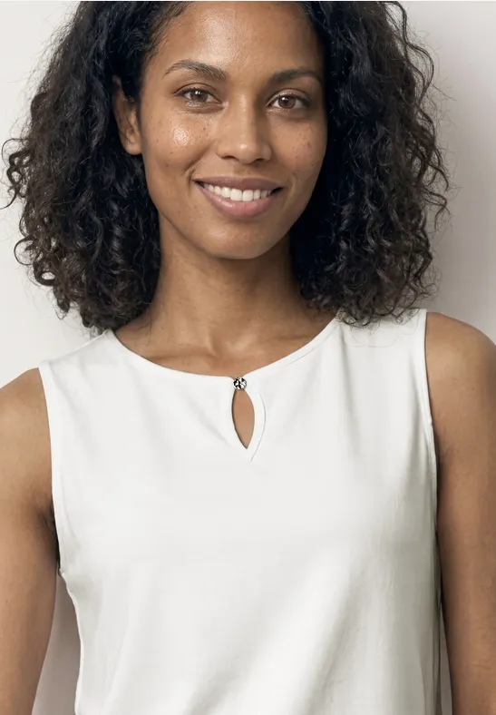 Vrouw met donker krullend haar, in mouwloze witte top met sleutelgatdetail, gezicht onscherp.