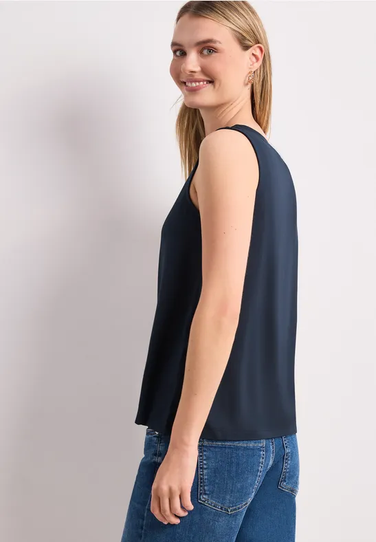 Een vrouw in een donkerblauw mouwloos topje en spijkerbroek, zijaanzicht tegen lichte achtergrond.