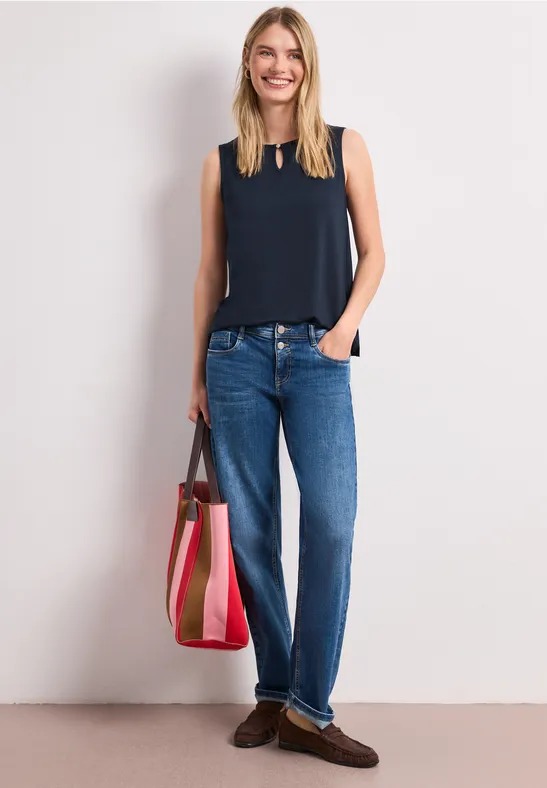 Vrouw met marine top zonder mouwen, blauwe jeans, bruine loafers en grote roze-beige gestreepte tas.