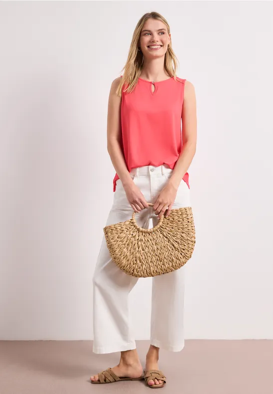Frau in pinkem Top und weißen Culottehose, trägt Basttasche und Sandalen vor neutralem Hintergrund.