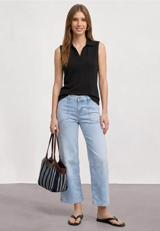 Frau in schwarzem Top, hellen Jeans, Flip-Flops, gestreifte Tasche, vor neutralem Hintergrund.