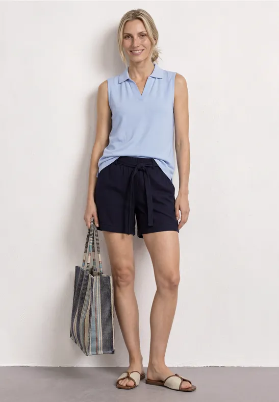 Vrouw in mouwloze lichtblauwe polo, donkerblauwe shorts, gestreepte tas en sandalen voor witte achtergrond.