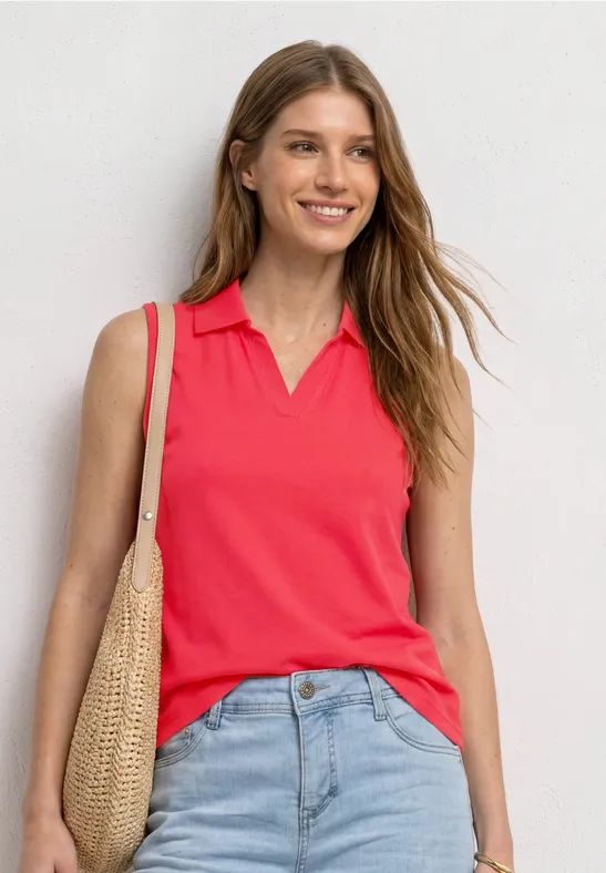 Femme devant un mur blanc, portant un top polo sans manches rouge, jean bleu et sac en paille.