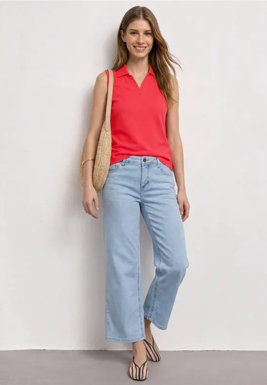 Femme en polo rouge sans manches, jean large bleu clair, sandales rayées et sac en paille devant un mur blanc.