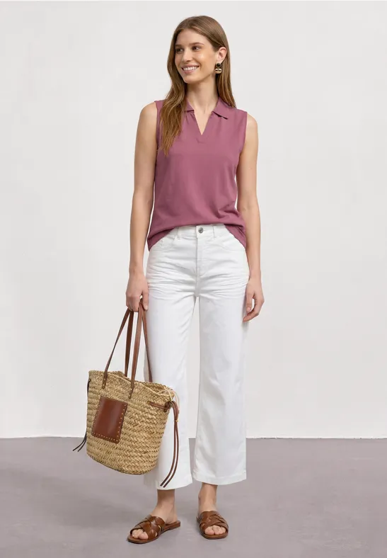 Femme en haut rose sans manches, pantalon blanc et sandales brunes avec un sac en paille tressée.