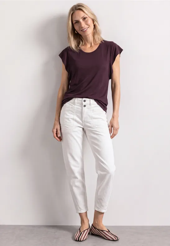 Eine Frau trägt ein locker sitzendes T-Shirt, weiße Hose mit Knopfleiste und Sandalen vor hellem Hintergrund.