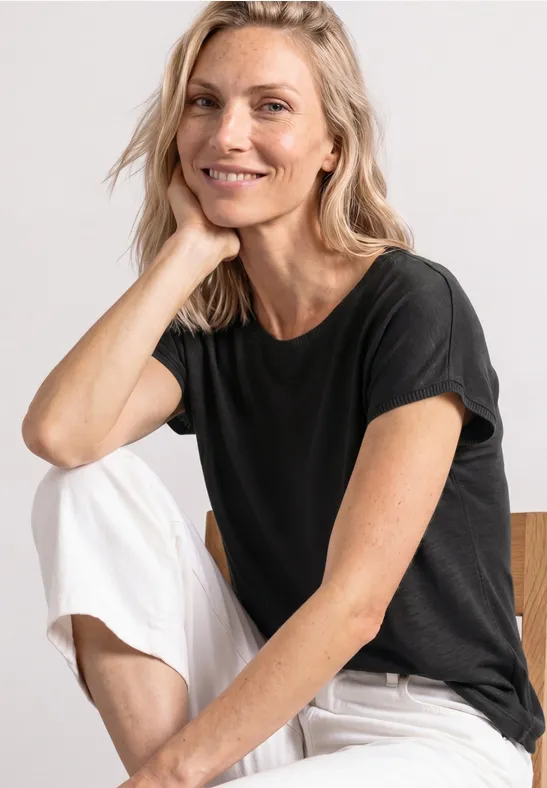 Vrouw zit in zwarte T-shirt en witte broek voor een neutrale achtergrond.