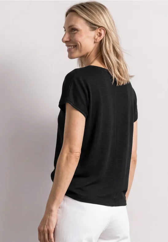 Vrouw van achteren in een zwart T-shirt en witte broek, licht zijaanzicht, neutrale achtergrond.