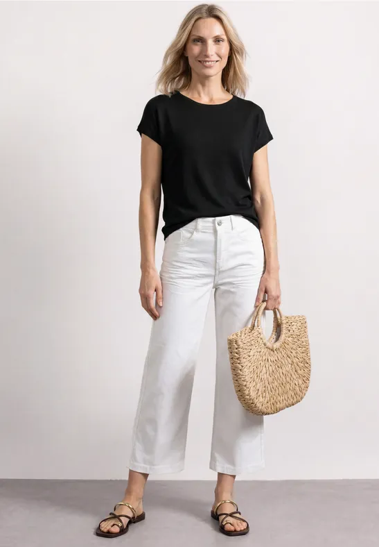 Vrouw in zwart t-shirt, witte jeans en sandalen met strooien tas voor een lichte achtergrond.