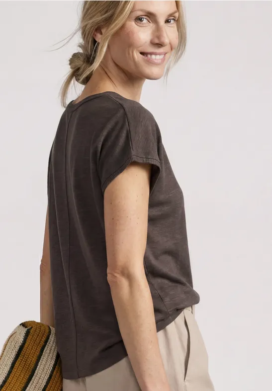 Profil d'une femme en t-shirt foncé et pantalon clair, cheveux en tresse, fond uni.