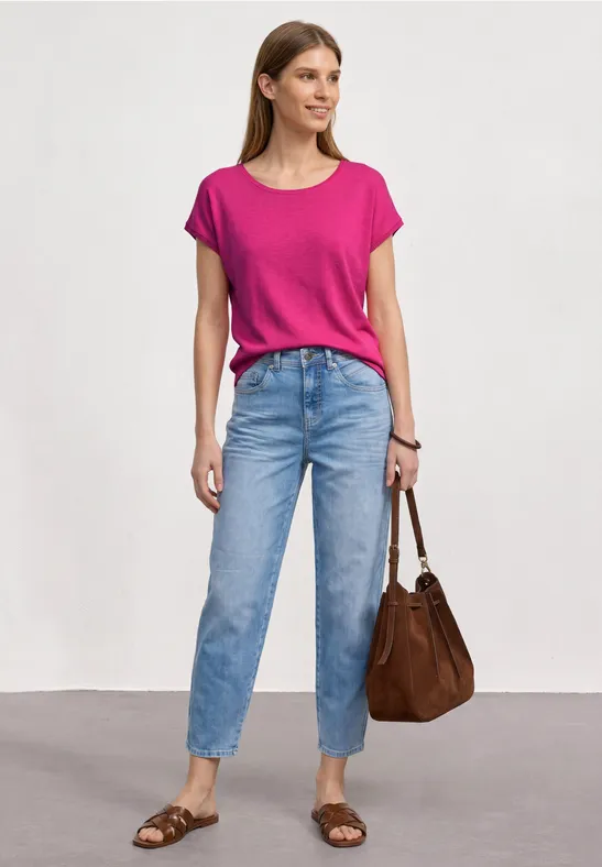 Vrouw in een roze t-shirt, blauwe jeans, bruine tas en sandalen voor een neutrale achtergrond.