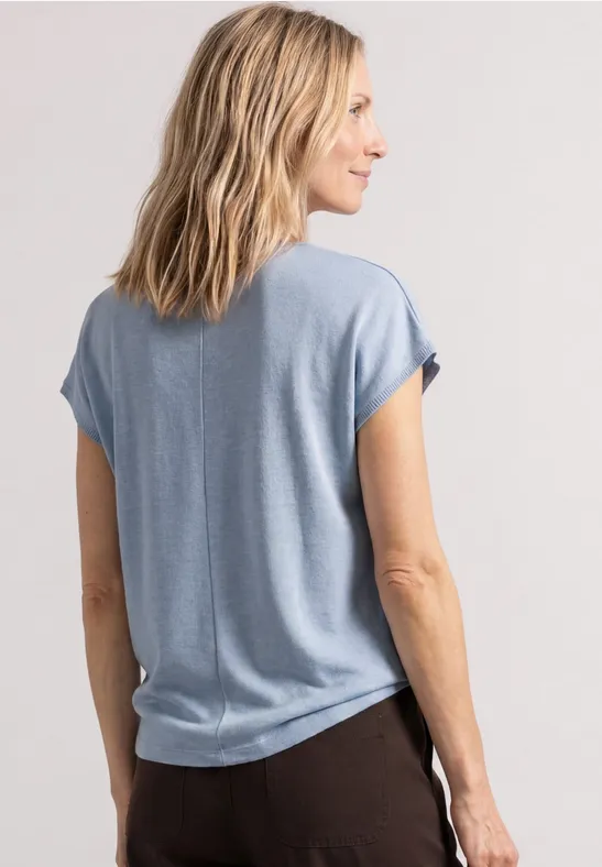 Eine Frau von hinten in einem hellblauen T-Shirt und dunklen Hosen, stehend und seitlich blickend.