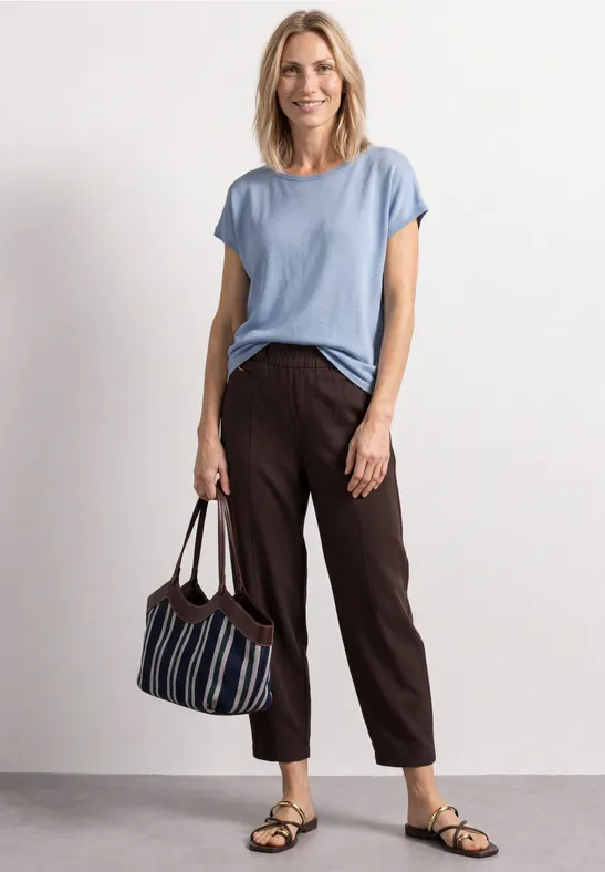 Frau trägt hellblaues T-Shirt, braune Hose, Sandalen und hält eine gestreifte Tasche.