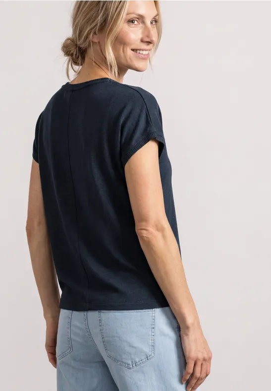 Eine Frau im Profil dreht sich lächelnd um, trägt dunkelblaues T-Shirt und helle Jeans.