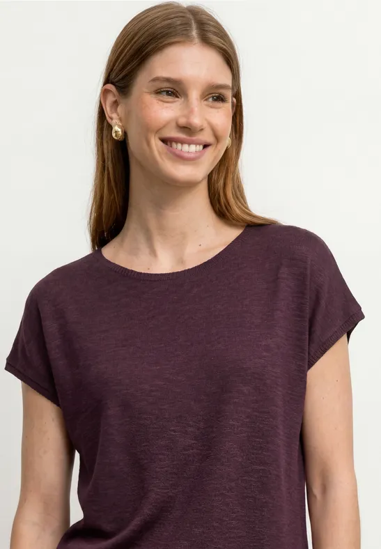 Frau mit langen Haaren trägt ein schlichtes, dunkelviolettes T-Shirt und goldene Ohrringe vor weißem Hintergrund.