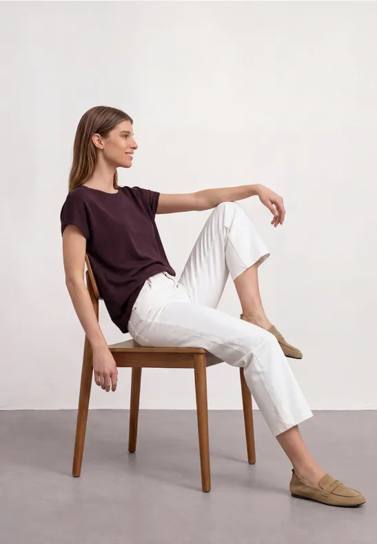 Junge Frau sitzt auf einem Stuhl, trägt ein bordeauxfarbenes T-Shirt, weiße Hose und beige Slipper.