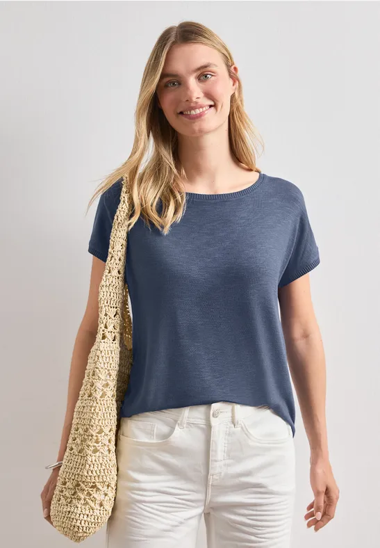 Frau in blauem T-Shirt und weißer Hose mit Basttasche über der Schulter.