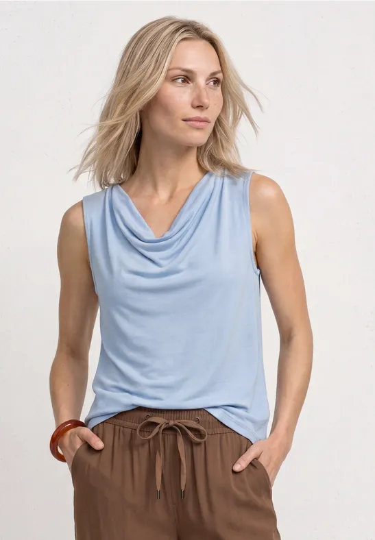 Frau mit hellblauem ärmellosem Top und braunen Hosen vor hellem Hintergrund.