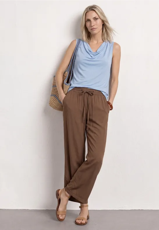 Frau lehnt in lässigem Outfit an weißer Wand: hellblaues Top, braune Hose, Sandalen und Strohtasche.