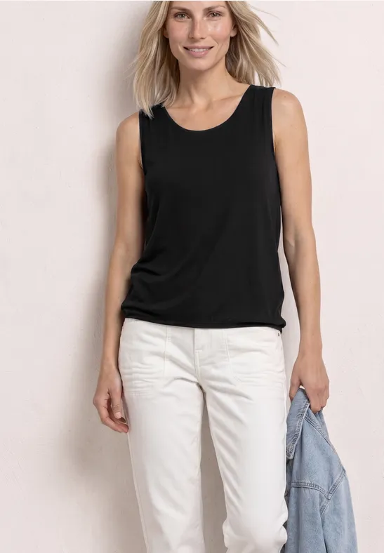 Frau in schwarzem ärmellosem Top und weißen Hosen, hält Jeansjacke in der Hand, vor hellem Hintergrund.