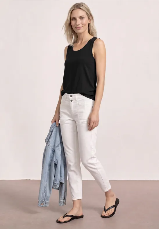 Eine Frau trägt ein schwarzes Top, weiße Jeans, schwarze Flip-Flops und hält eine hellblaue Jeansjacke.