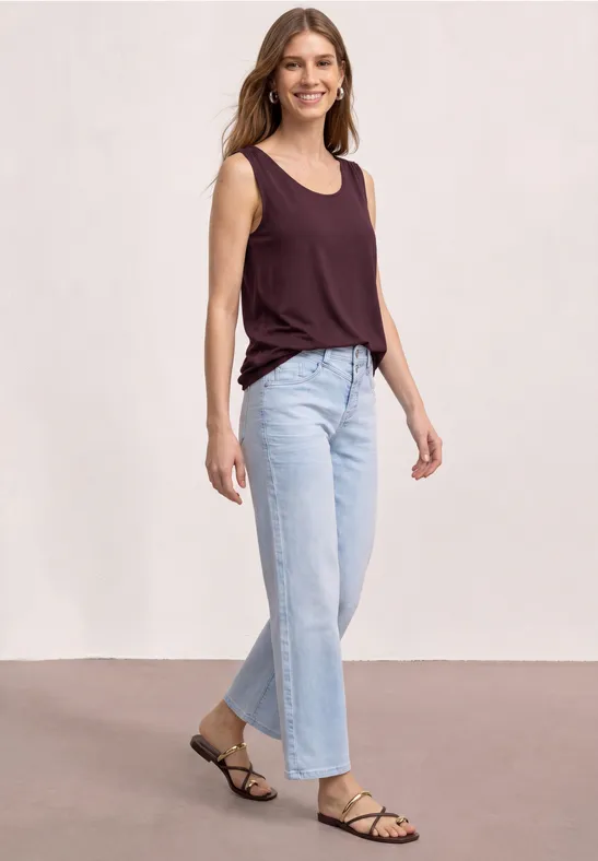 Frau in ärmellosem bordeauxfarbenen Top, weiten hellen Jeans und schlichten Riemchensandalen.