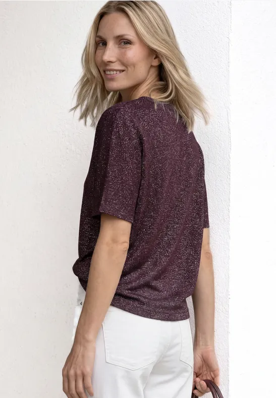 Eine von hinten gezeigte Person trägt ein glitzerndes T-Shirt und weiße Jeans vor weißem Hintergrund.
