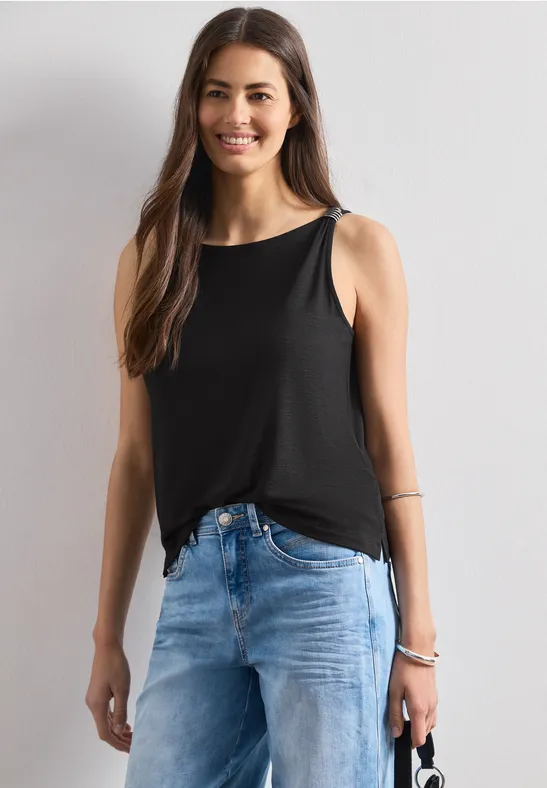 Een vrouw draagt een zwart mouwloos topje en blauwe jeans, ze houdt een klein tasje in haar linkerhand.