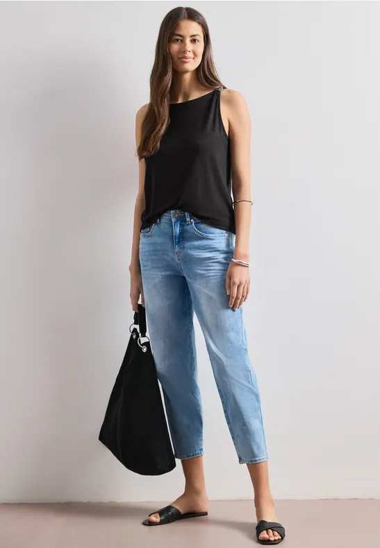 Vrouw in zwarte mouwloze top, lichtblauwe jeans en zwarte sandalen, met zwarte handtas.