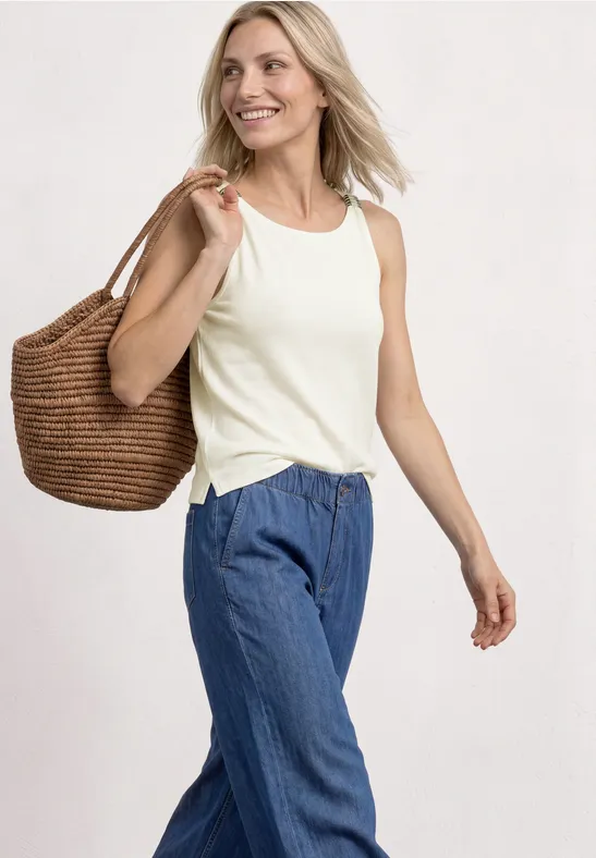 Vrouw met wit mouwloos topje, blauwe broek en strooien tas over haar schouder, lopend voor een lichte achtergrond.