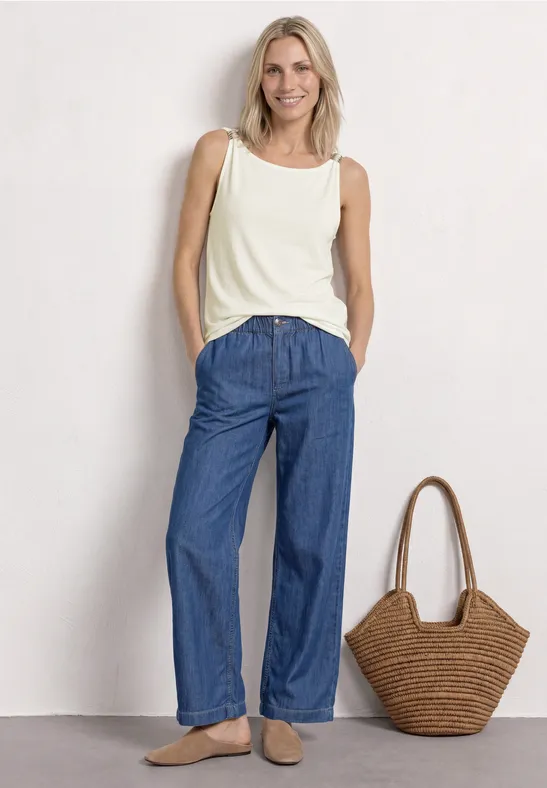 Vrouw in witte tanktop en wijde jeans, staand naast een rieten tas.