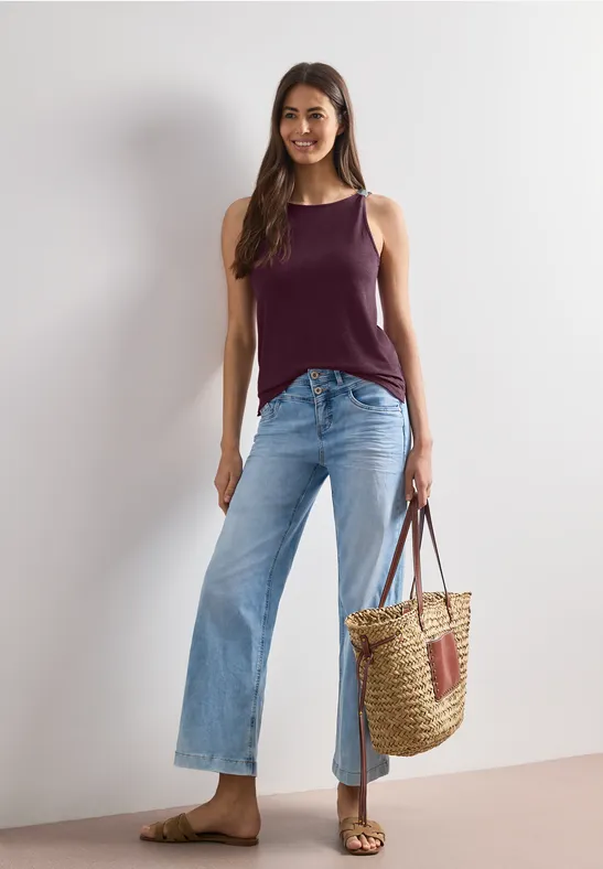 Vrouw voor neutrale achtergrond in casual outfit: donker topje, wijde spijkerbroek, sandalen en strooien tas.