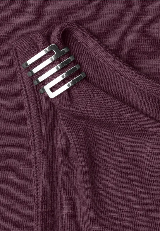 Close-up van een bordeauxrode shirt met borstzak en een verzilverd metalen clip erop vastgemaakt.