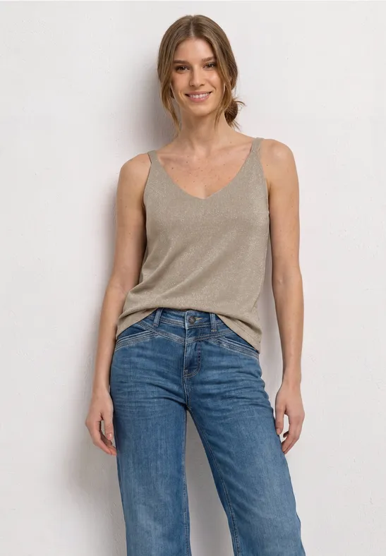 Frau in beigem Top und blauen Jeans vor weißer Wand.