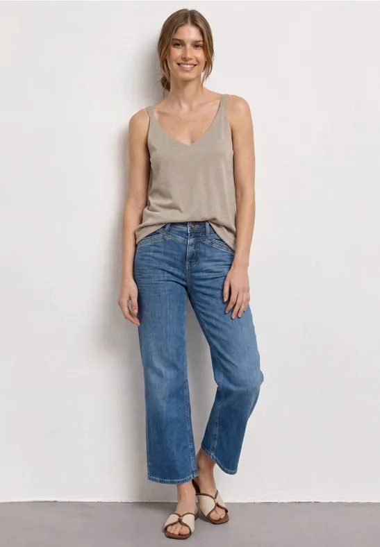 Frau in beigem Top, blauen, weit geschnittenen Cropped-Jeans und Sandalen vor weißer Wand.