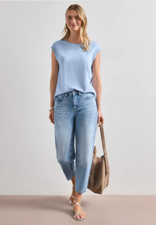 Frau in hellblauem Shirt, lockeren Jeans und Sandalen, trägt Shopper-Tasche vor neutralem Hintergrund