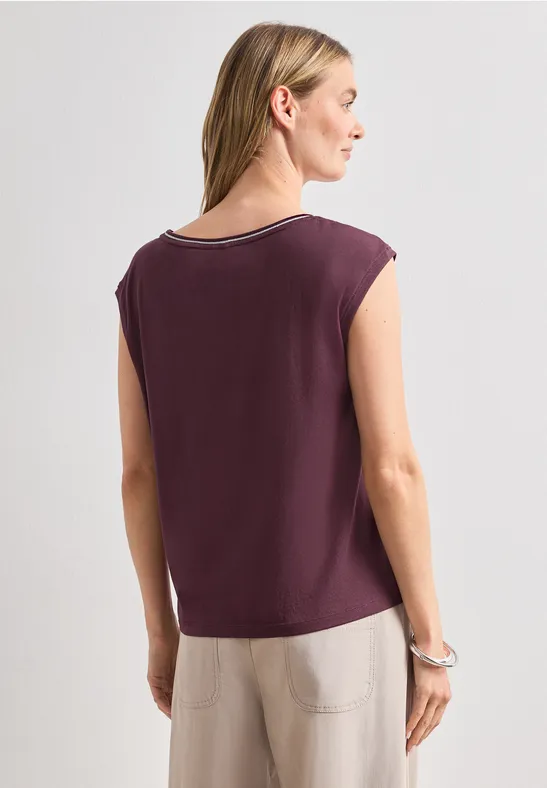 Vrouw van achteren in een donkerpaars mouwloos shirt en een beige broek tegen een neutrale achtergrond.