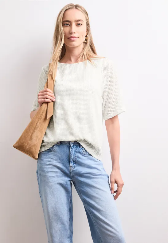 Frau mit beigem T-Shirt, hellblauen Jeans und brauner Schultertasche vor heller Wand stehend.