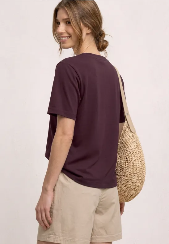 Frau in violettem T-Shirt, beigen Shorts und runder Strohtasche, Rückenansicht.