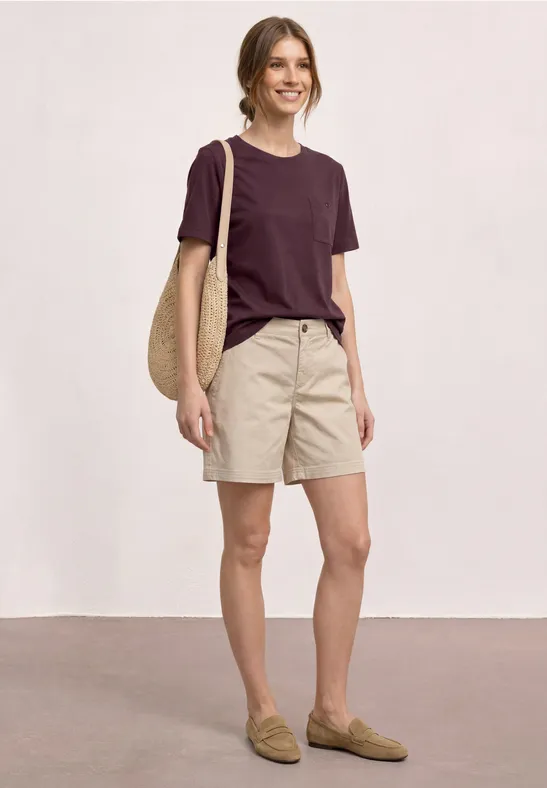 Person in legerer Kleidung: dunkelrotes T-Shirt, beige Shorts, Loafers und Korbtasche.