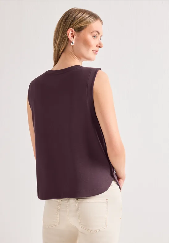 Femme de dos portant un top sans manches violet sombre et un pantalon clair.