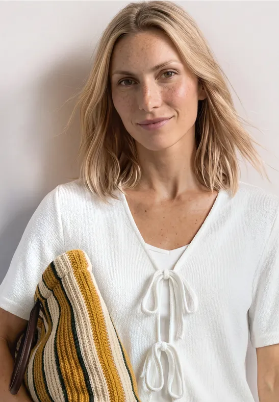 Eine Person trägt eine weiße Bluse mit Zierschleifen und hält ein gestreiftes Strickteil.