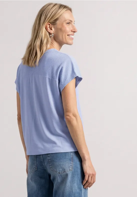 Eine Frau mit blonden Haaren trägt ein hellblaues T-Shirt und Jeans, im Profil, vor neutralem Hintergrund.