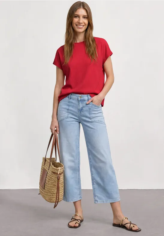 Vrouw in rood T-shirt, lichtblauwe cropped jeans, zwarte sandalen en strooien shopper op neutrale achtergrond.