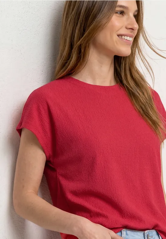 Vrouw steunt tegen een witte muur, draagt een rood T-shirt met korte mouwen en spijkerbroek.